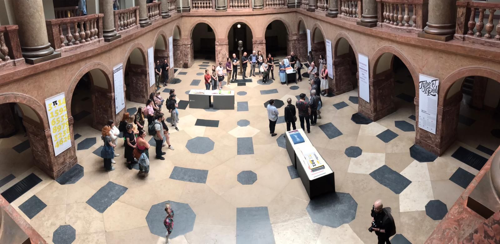 Widok z górnej galerii na hol Collegium Maius, po którym przechadzają się różne osoby, oglądając wystawione ekspozycje.