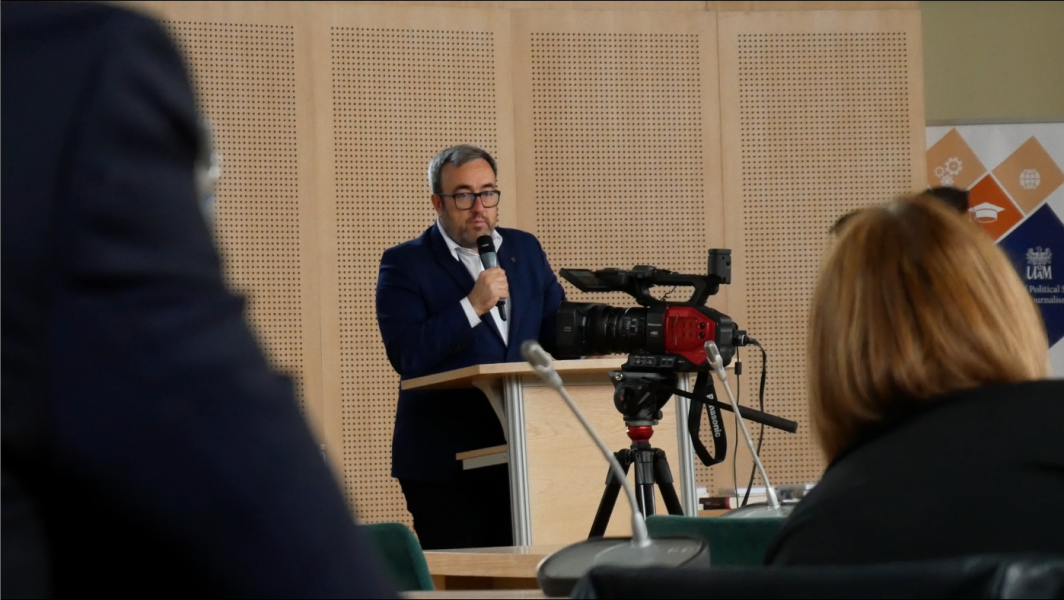 Szpakowaty mężczyzna w garniturze i okularach przemawia do mikrofonu przy mównicy, przed nim stoi kamera. Zdjęcie zrobione zza pleców publiczności.