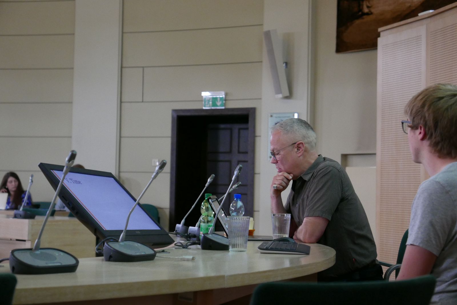 Prof. Anno Mungen siedzi za stołem prezydialnym w Salonie Mickiewicza, wygłaszając wykład do mikrofonu. Patrzy w ekran tabletu. W tle widać słuchaczy.