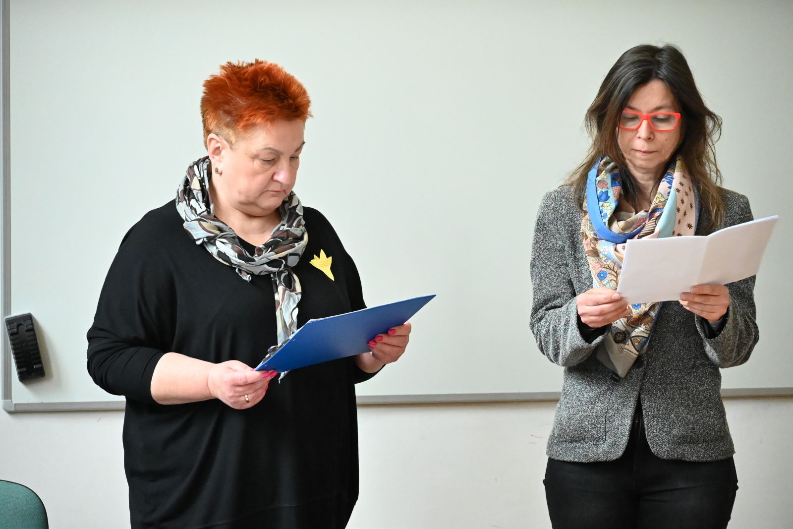 Dwie kobiety stoją za stołem, wpatrują się w teksty, które trzymają w dłoniach.