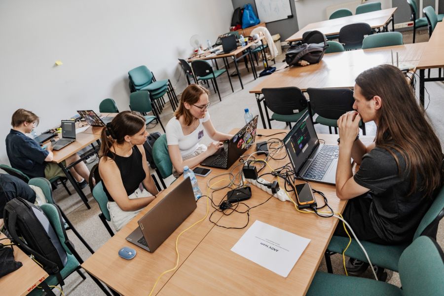 Młode osoby siedzą przy stołach z laptopami i programują.