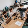 Młode osoby siedzą przy stołach z laptopami i programują.