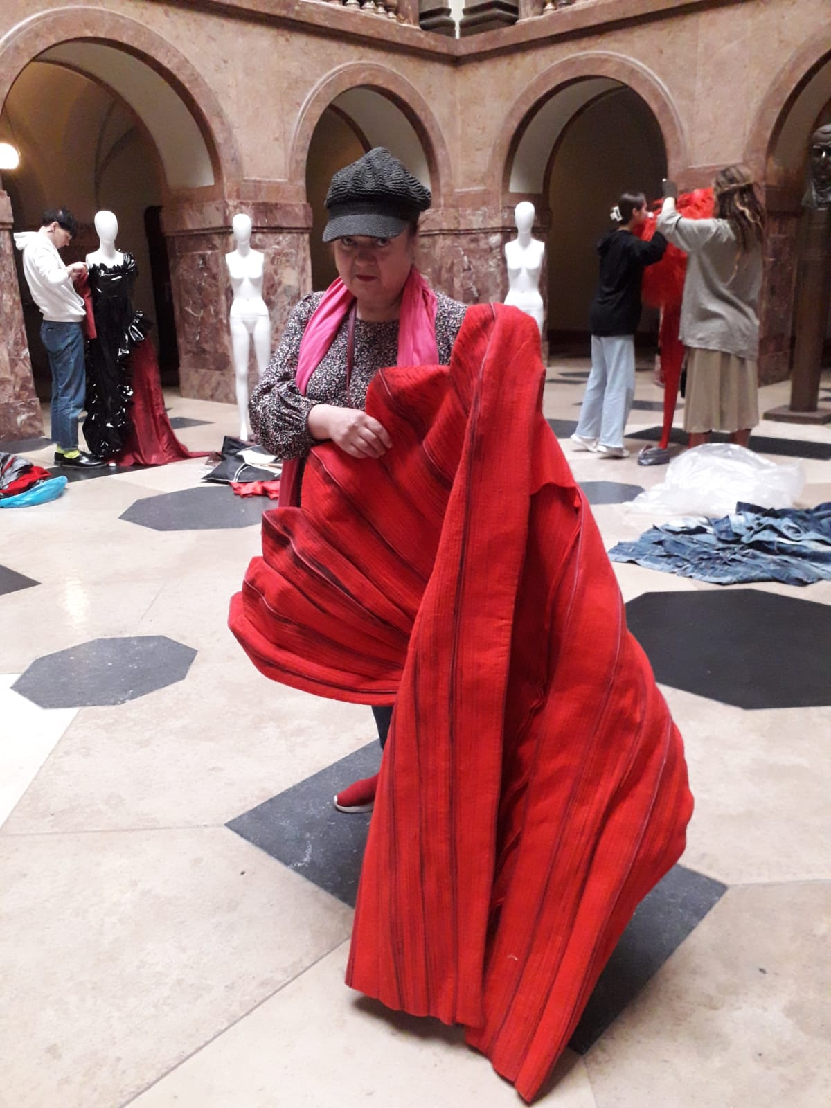 Kobieta w średnim wieku prezentuje czerwoną tkaninę uszytą w kontrafałdy, za nią widać manekiny i osoby, które je ubierają.