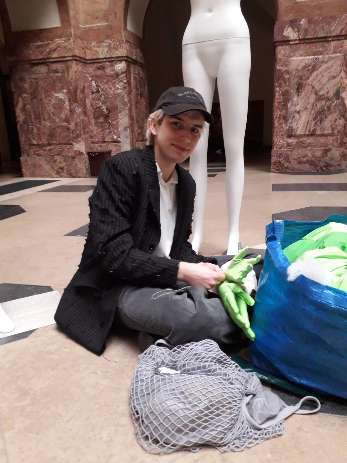 Młoda osoba siedzi po turecku w holu Collegium Maius, wyciąga tkaniny z toreb, patrzy w obiektyw. Za nią stoi nagi manekin.