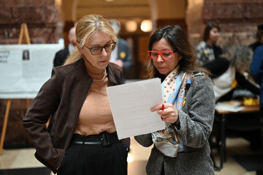 Zdjęcie przedstawia dwie kobiety w okularach, jedna trzyma kartkę papieru i pokazuje ją drugiej. W tle za nimi widać więcej osób.