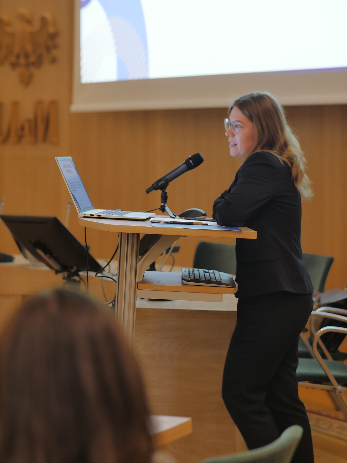 Młoda kobieta stoi przy mównicy, wygłaszając swój referat, za nią na ekranie wyświetla się prezentacja, przed nią na pulpicie leży laptop.