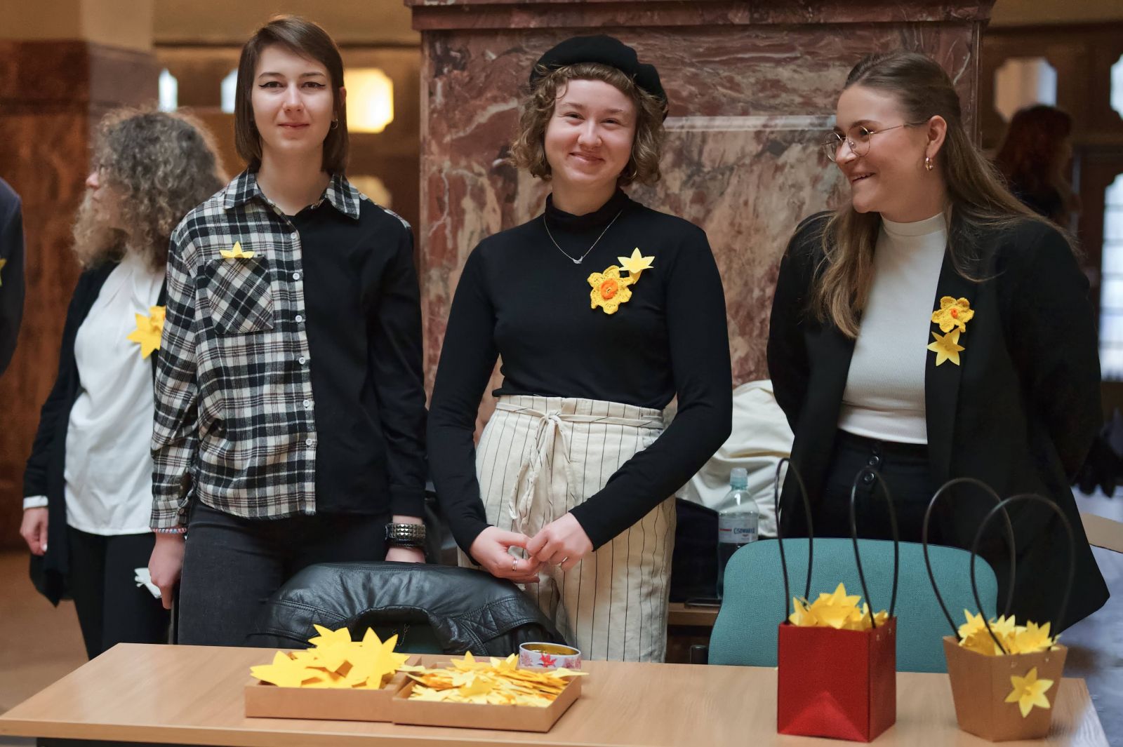 Trzy młode kobiety stoją za stołem, na którym leżą papierowe żonkile. Dwie z nich uśmiechają się do obiektywu, jedna patrzy w bok z uśmiechem. Na piersi mają przypięte papierowe i szydełkowe żółte żonkile. Za nimi widać jeszcze jedną kobietę, odwróconą bokiem do wida.