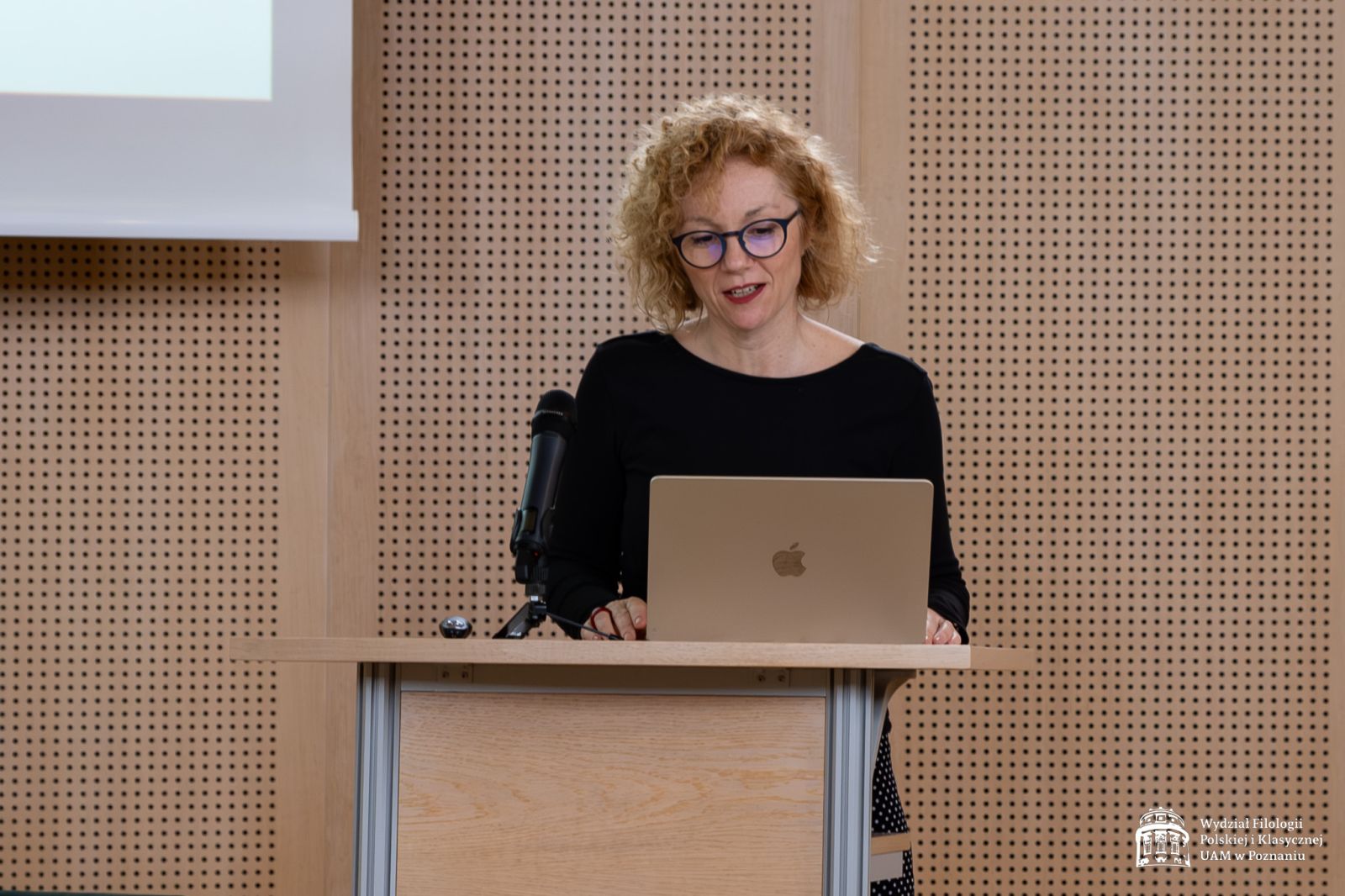 Blondwłosa kobieta w okularach stoi przy mównicy, patrzy w ekran laptopa, wygłaszając swoją prezentację.