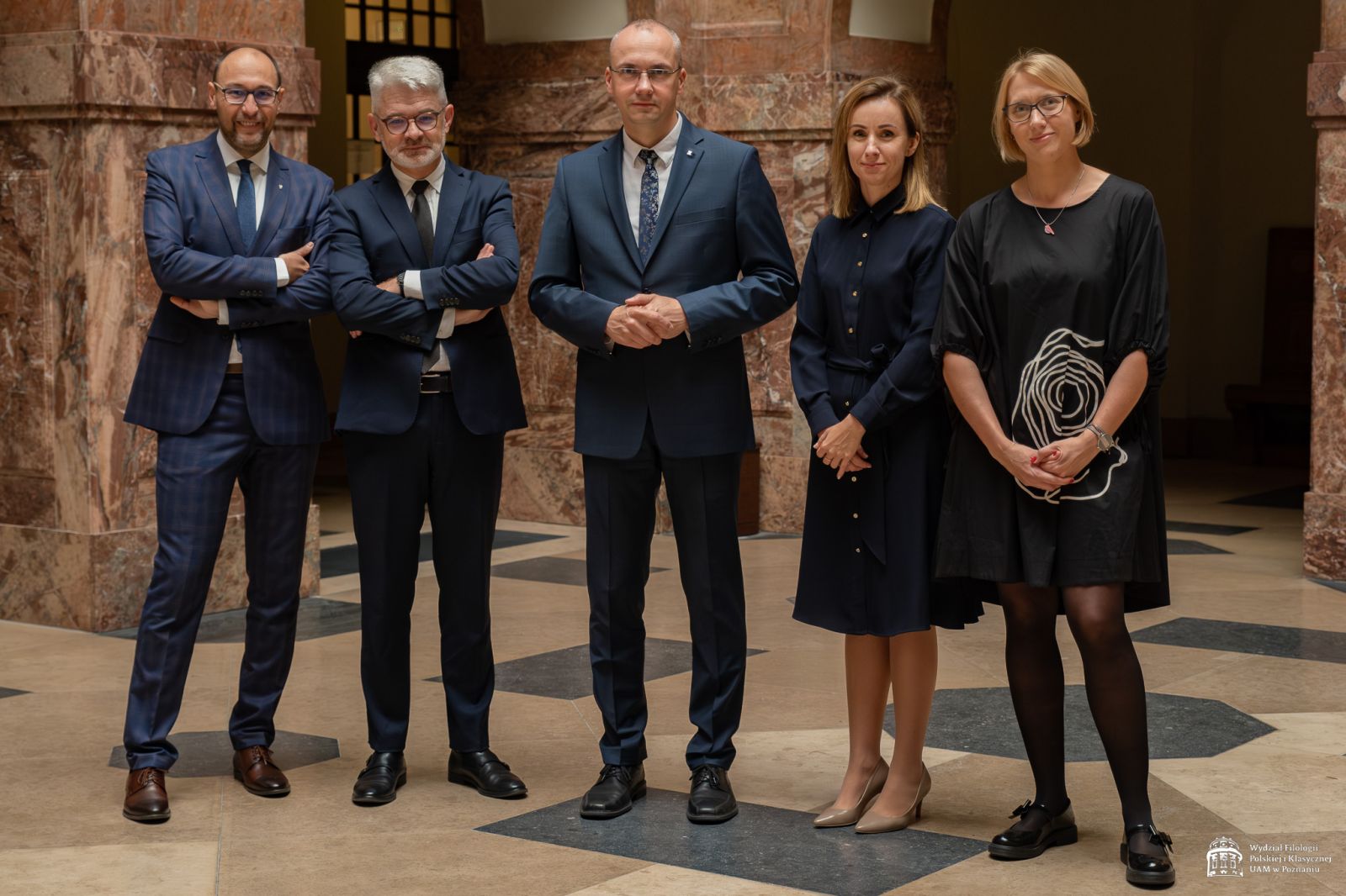 Na zdjęciu trzech mężczyzn i dwie kobiety stoi w holu głównym Collegium Maius, wszyscy są elegancko ubrani, uśmiechają się lekko do obiektywu.