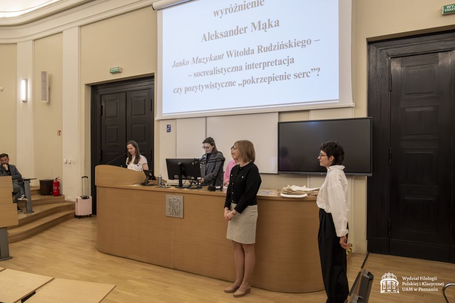 Prof. Paulina Michalska-Górecka stoi przy mikrofonie, prowadząc galę, obok niej za biurkiem stoi dr Aleksandra Nowak-Krystek, przed stołem stoją jeszcze dwie młode kobiety, na rzutniku wyświetla się tytuł nagrodzonej pracy.