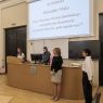 Prof. Paulina Michalska-Górecka stoi przy mikrofonie, prowadząc galę, obok niej za biurkiem stoi dr Aleksandra Nowak-Krystek, przed stołem stoją jeszcze dwie młode kobiety, na rzutniku wyświetla się tytuł nagrodzonej pracy.
