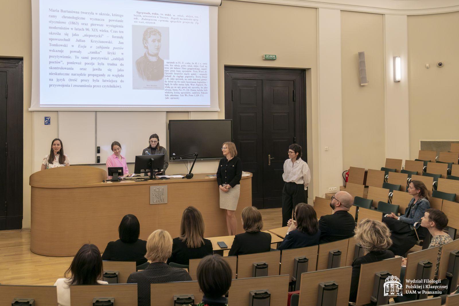 Zdjęcie stołu prezydialnego z prowadzącymi galę oraz część publiczności siedząca w rzędach.