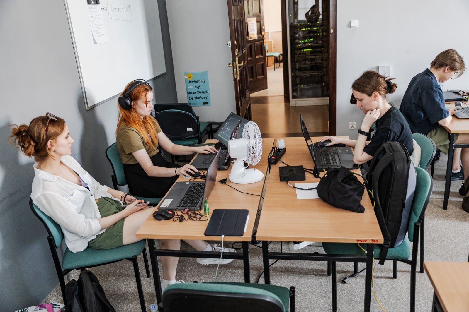 Młode osoby siedzą przy stołach z laptopami i programują.