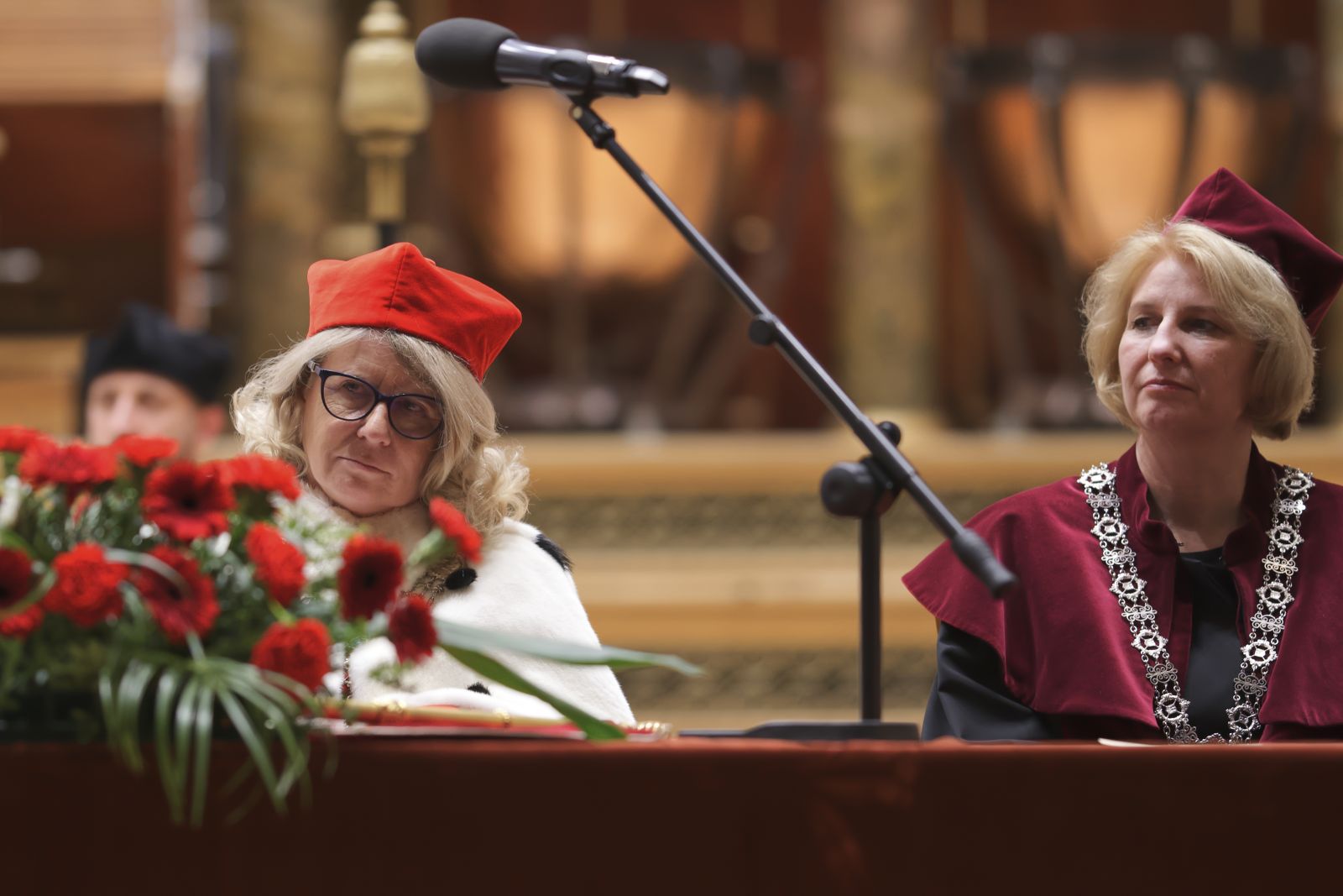 Rektor Bogumiła Kaniewska w czerwonej czapce i płaszczu gronostajowym oraz Prorektor Joanna Wójcik w bordowej czapce i płaszczu siedzą za stołem prezydialnym, na którym stoją czerwone kwiaty.
