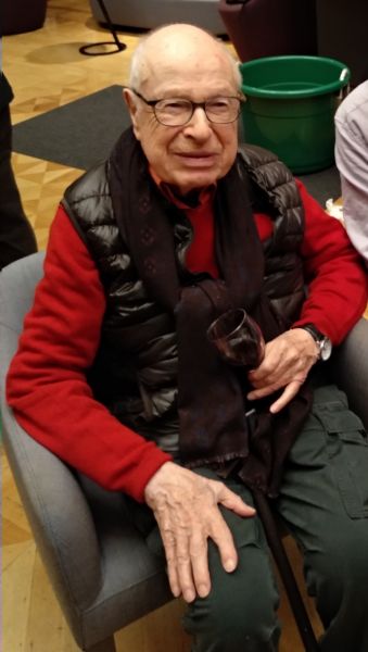 Starszy mężczyzna siedzi w fotelu, jest ubrany w czerwoną koszulę i czarną puchową kamizelkę, w dłoni trzyma kieliszek, na nosie ma okulary.