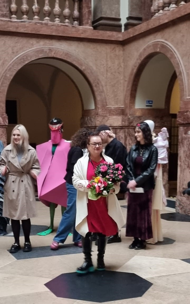 W holu głównym Collegium Maius stoi kilka osób, jedna z nich trzyma bukiet kwiatów i uśmiecha się. Za nimi widać manekiny ubrane w różnokolorowe szaty.
