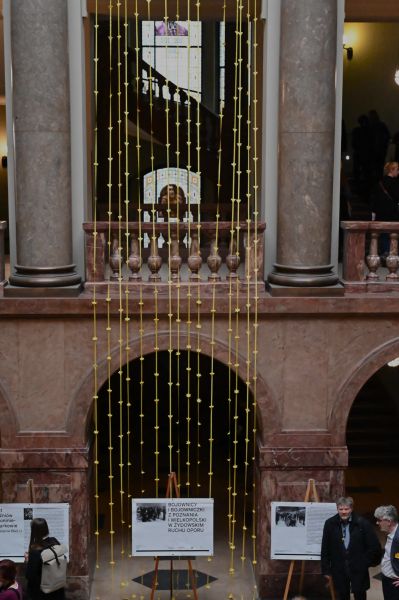 Zdjęcie części holu głównego Collegium Maius - z balustrady górnego piętra zwiesza się kurtyna zrobiona z żółtych wstążek, do której przyklejone są żółte papierowe żonkile. Na dole widać ludzi oglądających wystawę o wielkopolskich Żydach.
