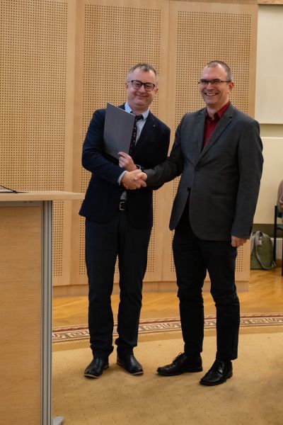 Dziekan WFPiK prof. Tomasz Mizerkiewicz wręcza nagrodę Rektor UAM prof. Mateuszowi Stróżyńskiemu, ściskając mu rękę. Obaj pozują do zdjęcia, szeroko się uśmiechając