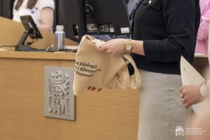 Zbliżenie na bawełnianą torbę ekologiczną z logo wydziału, trzymaną w dłoniach przez młodą kobietę. Za nią widać stół z dużym logo UAM.