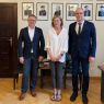 Troje osób - dwóch mężczyzn w garniturach i kobieta w jasnej koszuli - pozuje do wspólnego zdjęcia na tle ściany z portretami dawnych dziekanów WFPiK.