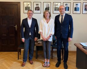 Troje osób - dwóch mężczyzn w garniturach i kobieta w jasnej koszuli - pozuje do wspólnego zdjęcia na tle ściany z portretami dawnych dziekanów WFPiK.