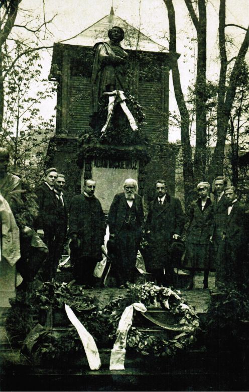 Czarno-białe zdjęcie przedstawiające grupę mężczyzn w ciemnych ubraniach, stojących przed pomnikiem Adama Mickiewicza