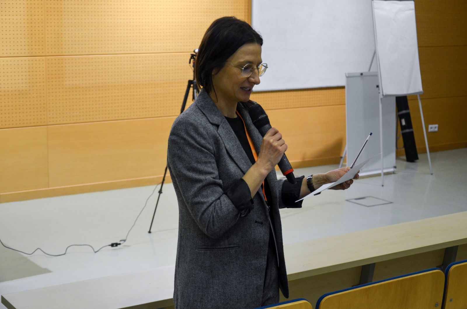 Prof. Barbara Sobczak czyta coś z kartki przez mikrofonu, stojąc przed widownią.  Za nią widać białą tablicę i flipcharty.