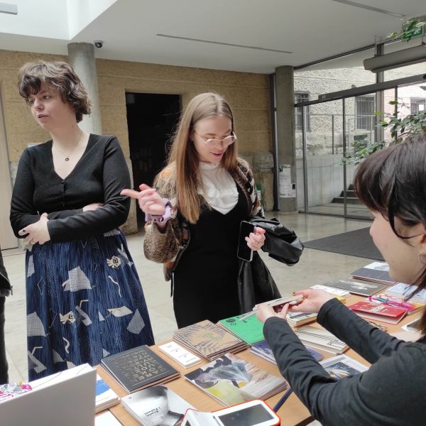 W holu biblioteki WFPiK przy stoisku z książkami stoją dwie młode osoby, jedna z nich ogląda prezentowaną przez młodą kobietę książkę.