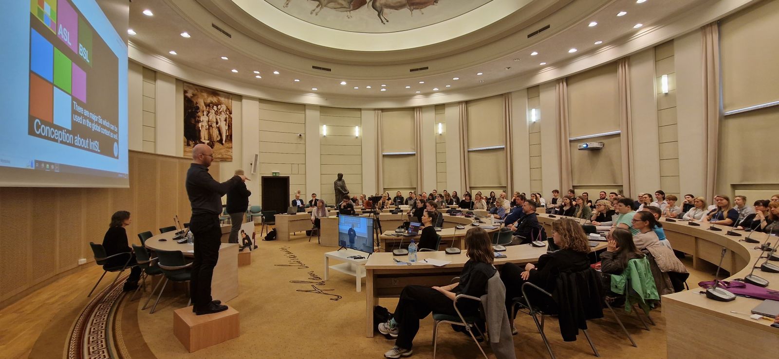 W Salonie Mickiewicza zgromadzeni są uczestnicy konferencji o języku migowym, przy stole prezydialnym na podwyższeniach stoją tłumacze na polski i uniwersalny język migowy.