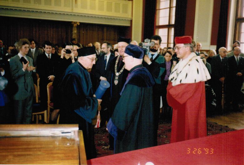 Kolorowe zdjęcie przedstawiające grupę osób ubranych w togi uniwersyteckie. Po lewej stronie rektor ubrany w czerwony płaszcz z gronostajową peleryną.