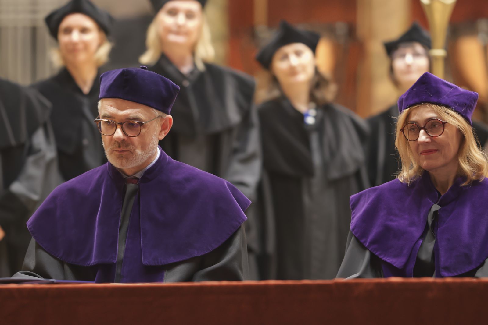 Prof. Jerzy Borowczyk i prof. Krystyna Pieniążek-Marković siedzą za stołem prezydialnym, oboje ubrani w fioletowe togi i czapki, za nimi stoją osoby w czarnych togach.