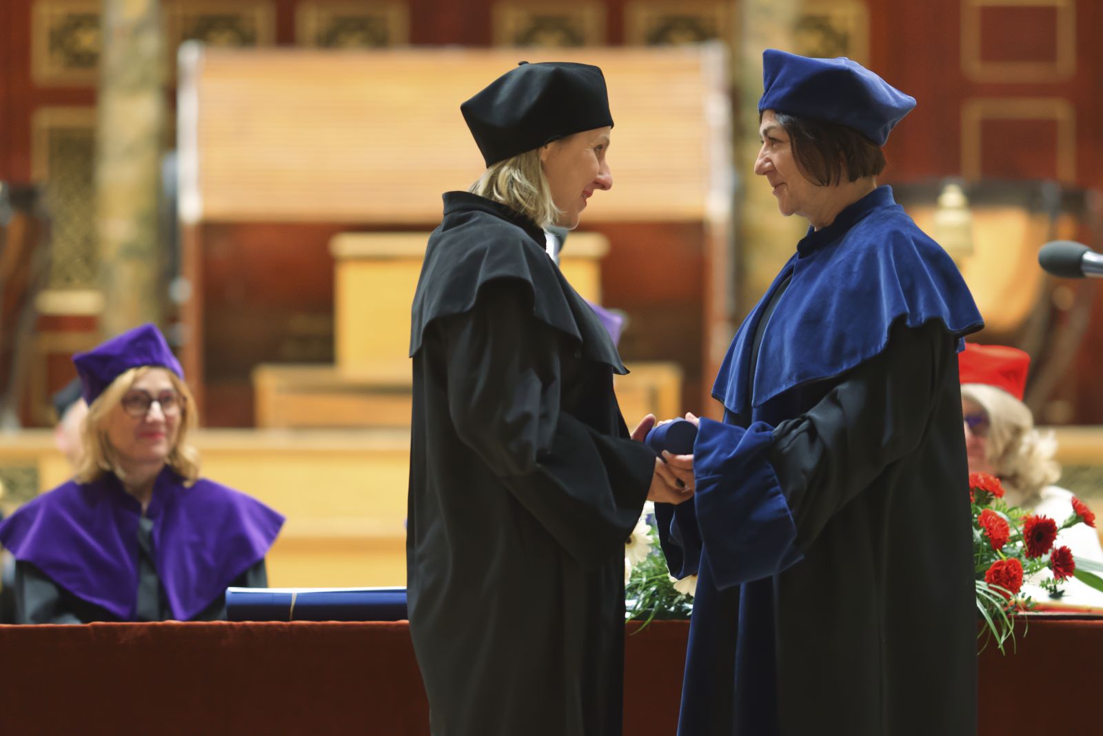 Dwie kobiety w togach, jedna wręcza drugiej dyplom, ściskając jej dłoń, za nimi widać stół prezydialny.