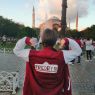 Zdjęcie kobiety w krótkich włosach i bordowej bluzie z białymi rękawami. Kobieta stoi tyłem, ma wzniesione ręce i palcami wskazującymi pokazuje na logotyp Fredry 10 na plecach swojej bluzy. Za nią widoczna Hagia Sophia w Stambule na tle zachodniego nieba. 