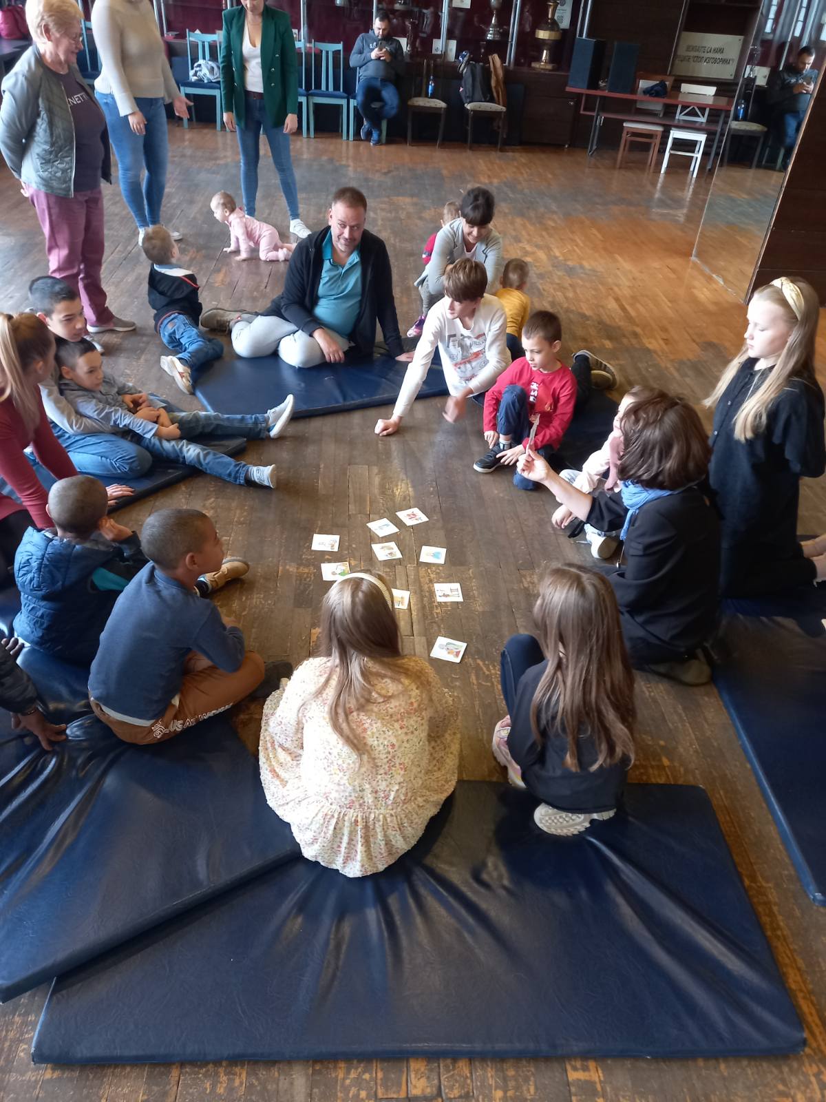 Na zdjęciu uczestnicy zajęć w Szkole Polskiej w Belgradzie kucają lub siedzą na drewnianej podłodze, oglądając kolorowe obrazki prezentowane przez prowadzącą.