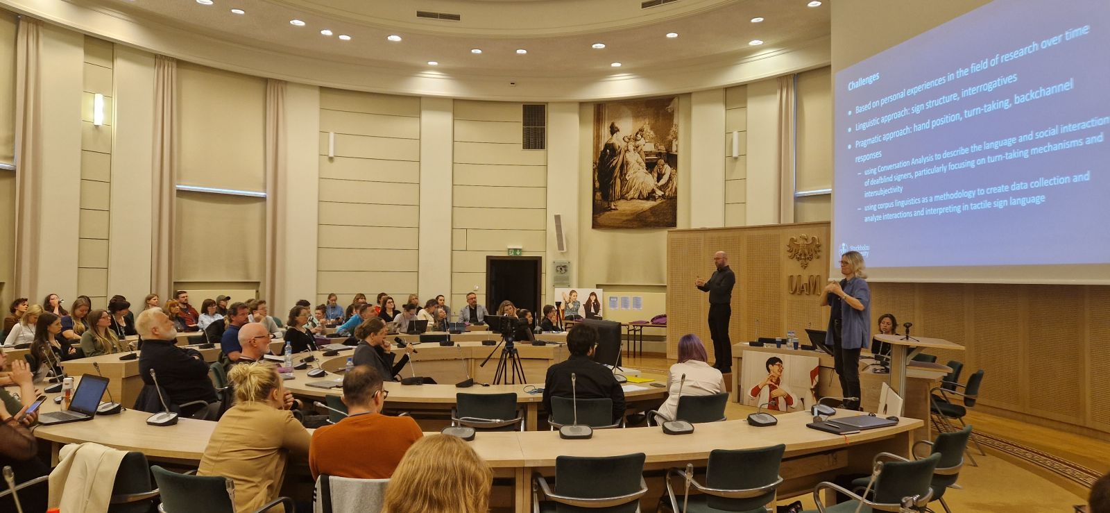 W Salonie Mickiewicza zgromadzeni są uczestnicy konferencji o języku migowym, przy stole prezydialnym na podwyższeniach stoją tłumacze na polski i uniwersalny język migowy.