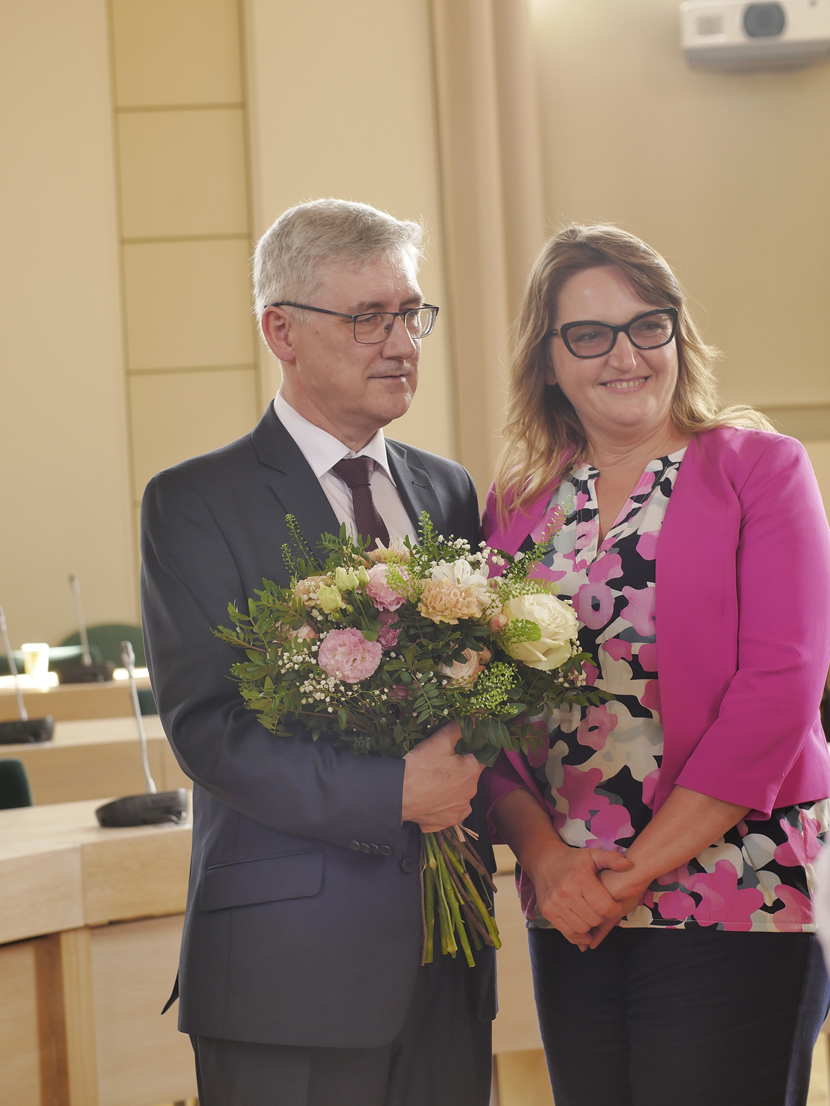 Prof. Jarosław Liberek pozuje do zdjęcia, trzymając bukiet kwiatów, obok niego stoi młoda roześmiana kobieta.