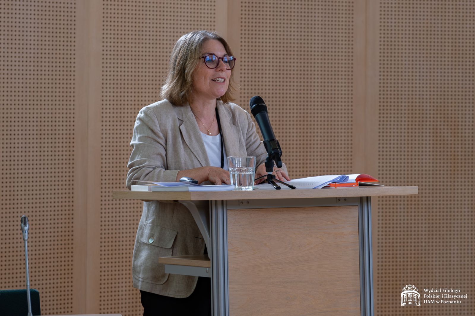 Kobieta o blond włosach i okularach, w szarej marynarce, stoi przy mównicy, wygłaszając swoje wystąpienie do mikrofonu.