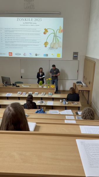 Zdjęcie z górnych rzędów ławek w sali Kuraszkiewicza, w których siedzą uczestnicy czytania, na dole wyświetla się prezentacja z programem wydarzenia, a przy mównicy stoi kobieta i mężczyzna, czytając tekst z kartki.