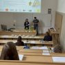 Zdjęcie z górnych rzędów ławek w sali Kuraszkiewicza, w których siedzą uczestnicy czytania, na dole wyświetla się prezentacja z programem wydarzenia, a przy mównicy stoi kobieta i mężczyzna, czytając tekst z kartki.