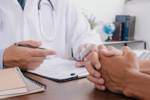 Zdjęcie dłoni spoczywających na biurku - lekarza ubranego w biały kitel, ze stetoskopem na szyi, który w dłoni trzyma długopis, a przed sobą ma wydrukowany formularz. Na przeciwko niego splecione dłonie pacjenta. W tle za biurkiem widać stojące na półce książki, globus i kwiaty.