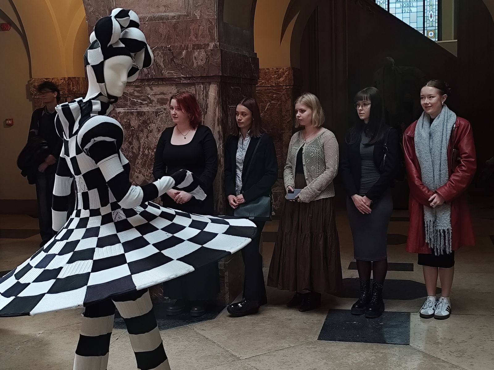 Studentki filologii polskiej podczas wernisażu, przed nimi stoi jeden z manekinów w czarno-białym kostiumie w kratkę.