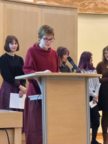 Przy mównicy w Salonie Mickiewicza stoi uczennica i odczytuje swoje wystąpienie, za nią stoją inni uczniowie i uczennice.