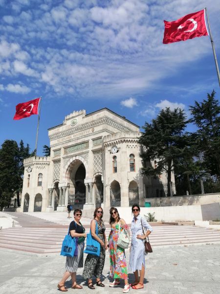 Cztery kobiety stoją na placu, za nimi biały budynek w stylu orientalnym, nad którym powiewa flaga turecka. 