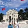 Cztery kobiety stoją na placu, za nimi biały budynek w stylu orientalnym, nad którym powiewa flaga turecka. 