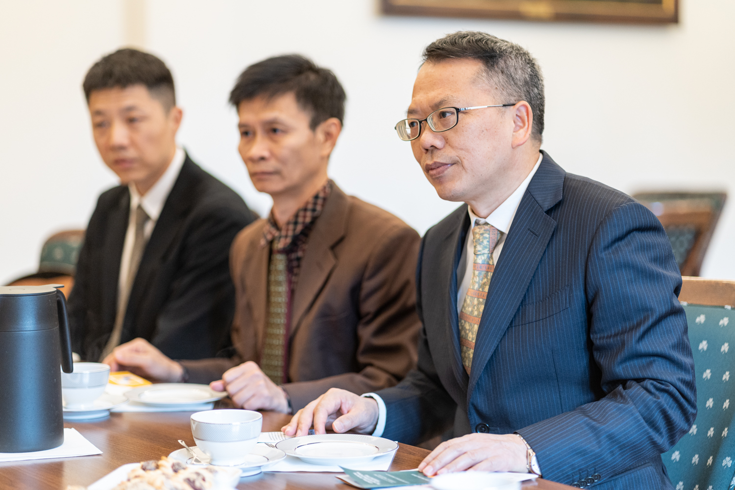 Trzech gości z Chin siedzi przy stole, uczestnicząc w rozmowie. Są ubrani w garnitury i krawaty.