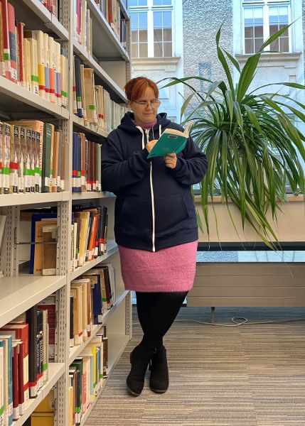 Dr Magdalena Baer stoi oparta o regał biblioteczny, w ręce trzyma otwartą książkę, którą czyta za pomocą lupki zawieszonej na łańcuszki na szyi.