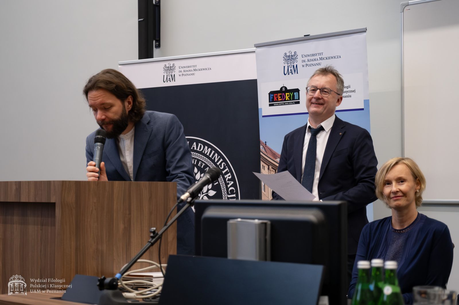 Dr Krzysztof Hoffmann przemawia przy mównicy do mikrofonu, pochylony nad pulpitem, za nim stoi uśmiechnięty dziekan WFPiK, obok siedzi prof. Katarzyna Klafkowska-Waśniowska.