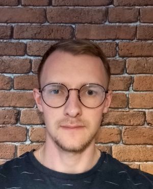 Zdjęcie młodego mężczyzny w okrągłych okularach i krótkich blond włosach, pozującego na tle ceglanej ściany.