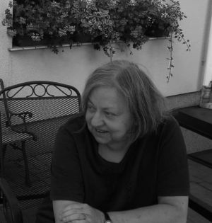 Czarno-białe zdjęcie starszej kobiety, siedzącej na ogrodowym krześle, za którym widać kwiaty doniczkowe na parapecie okna. Kobieta uśmiecha się, rozmawiając z osobą spoza kadru.