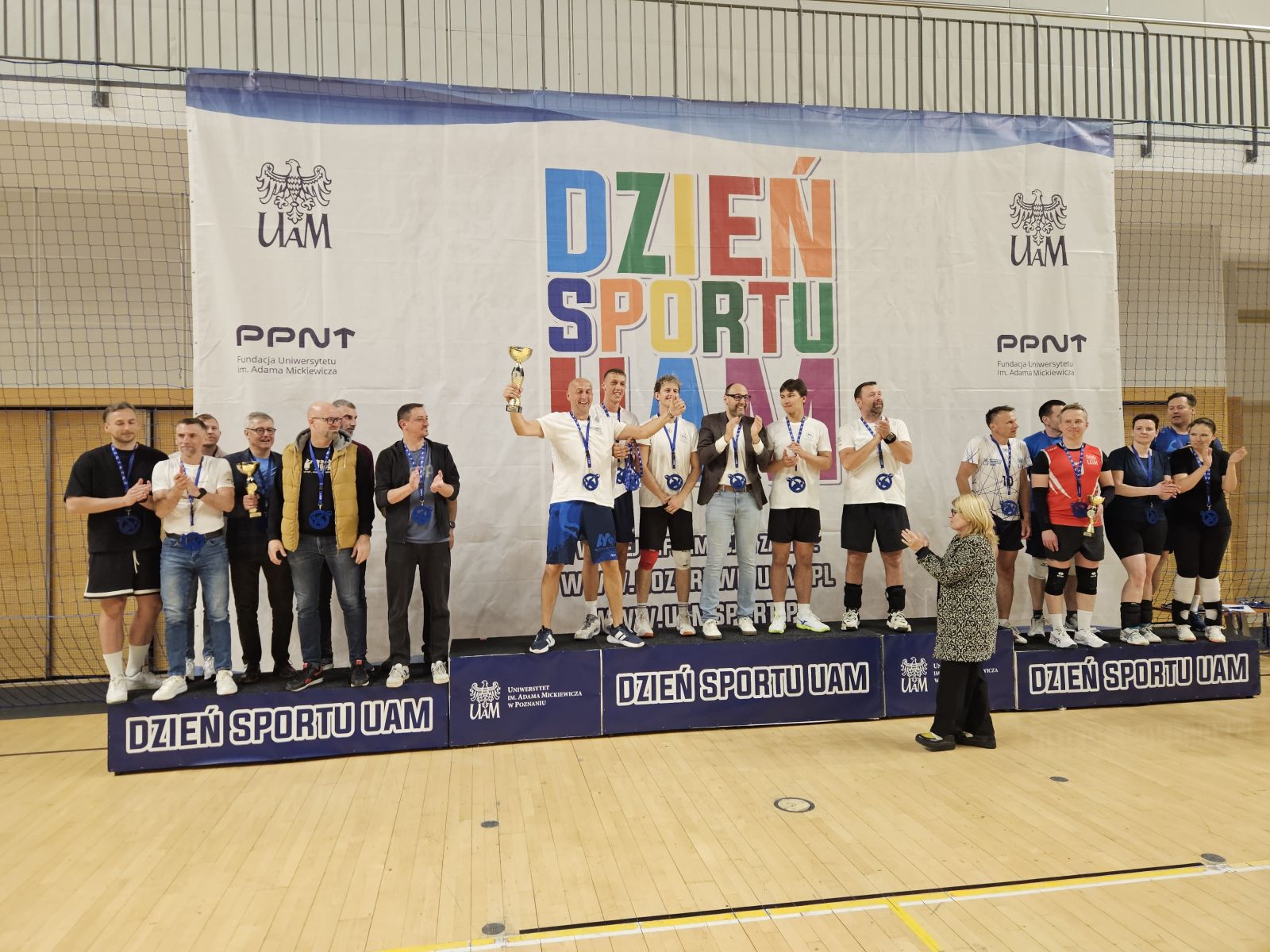 Drużyna siatkarska stoi na szczycie podium, ciesząc się z pucharu.